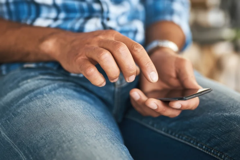 Man using smartphone while sitting, hands on device, wearing blue plaid shirt and jeans