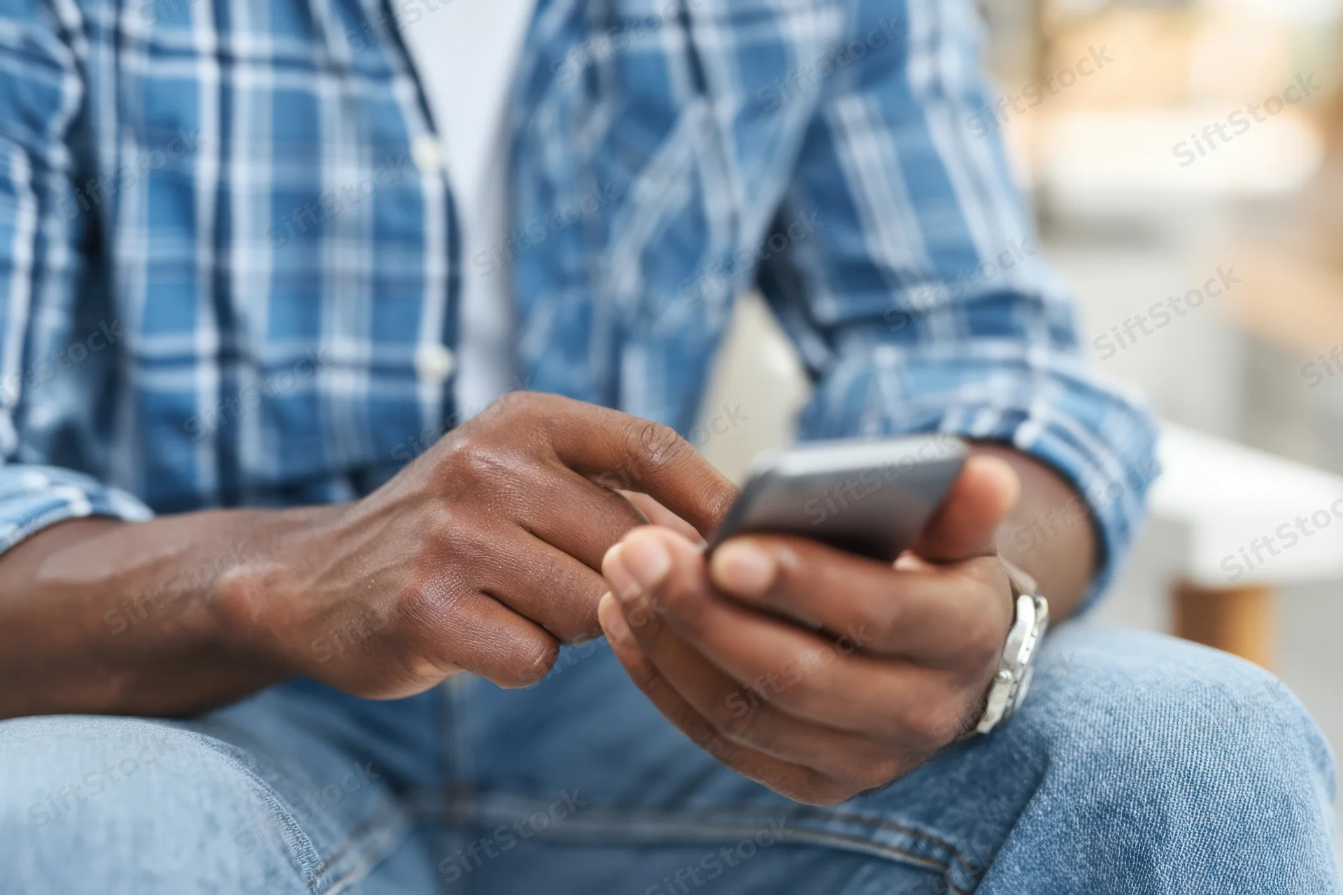 Man texting on smartphone while seated, wearing blue plaid shirt and jeans