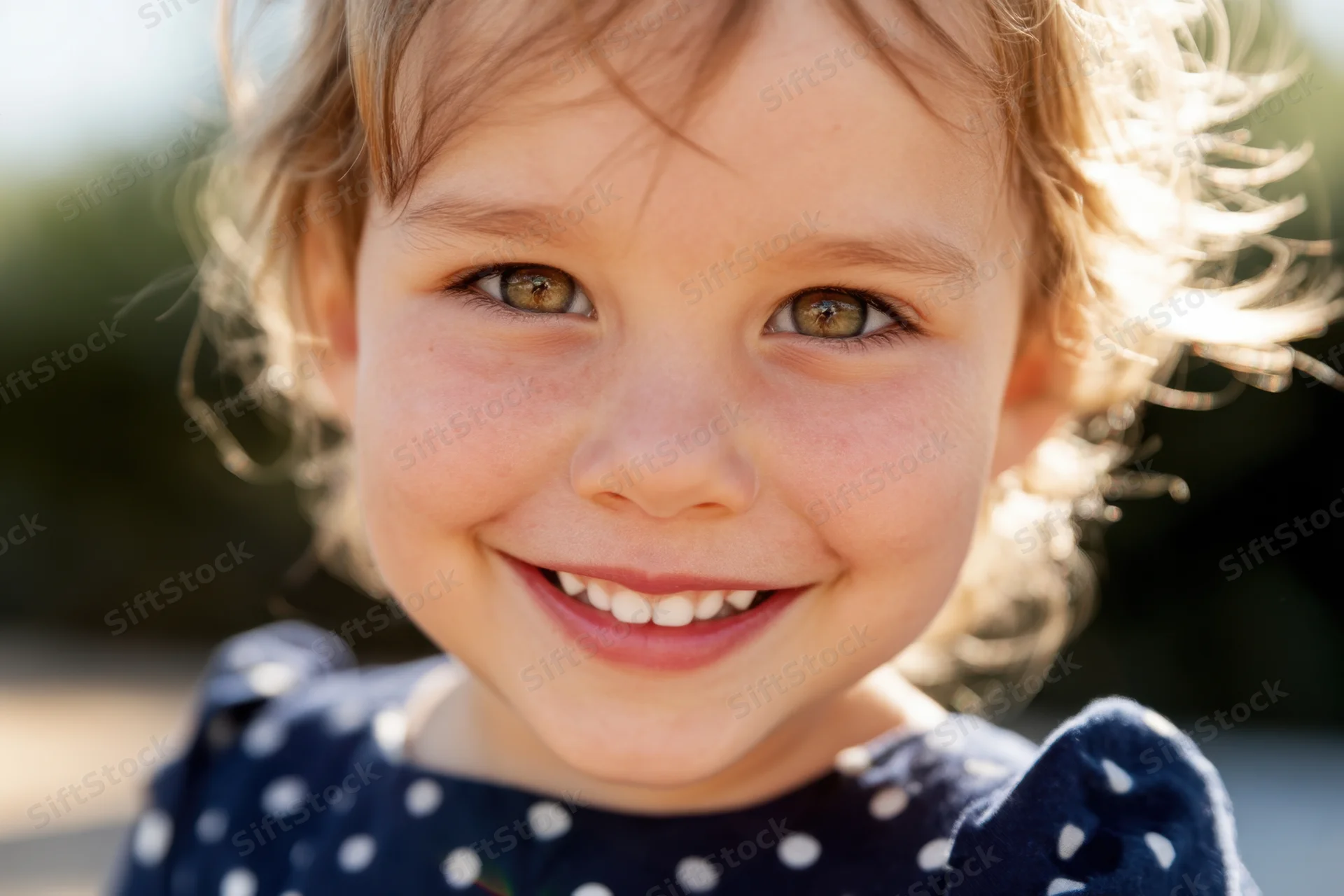 Close-up of a smiling toddler girl with light brown curly hair and hazel eyes wearing a navy polka dot dress outdoors in sunlight