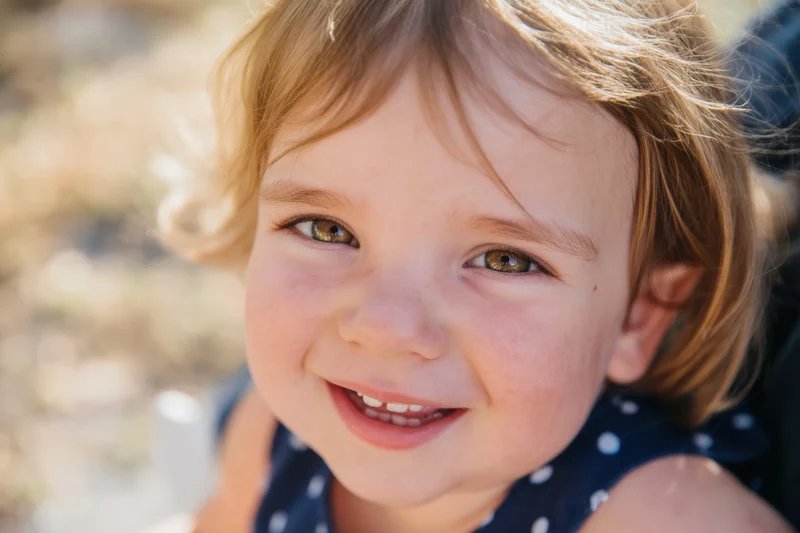 Smiling toddler with light brown hair and hazel eyes in a blue polka dot dress outdoors