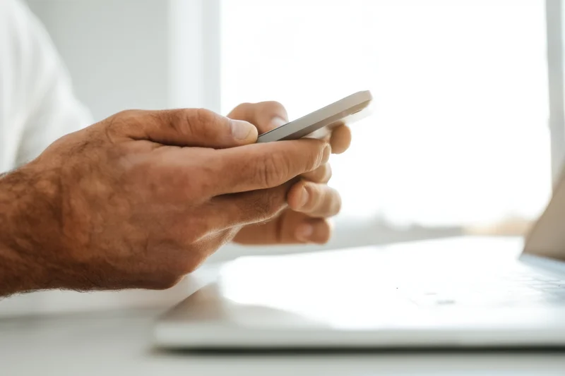 Close-up of male hands holding smartphone above laptop on desk