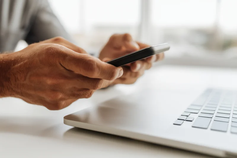 Hands holding smartphone above laptop on desk