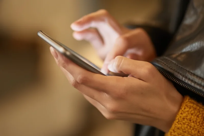 Hands typing on a smartphone held in a leather jacket sleeve with a yellow knit cuff visible