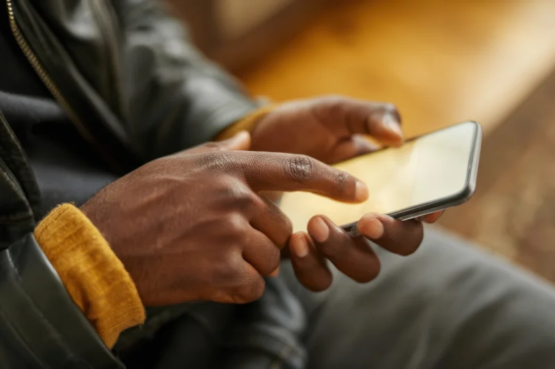 Man’s hands holding smartphone with finger poised over screen while wearing leather jacket and yellow cuff