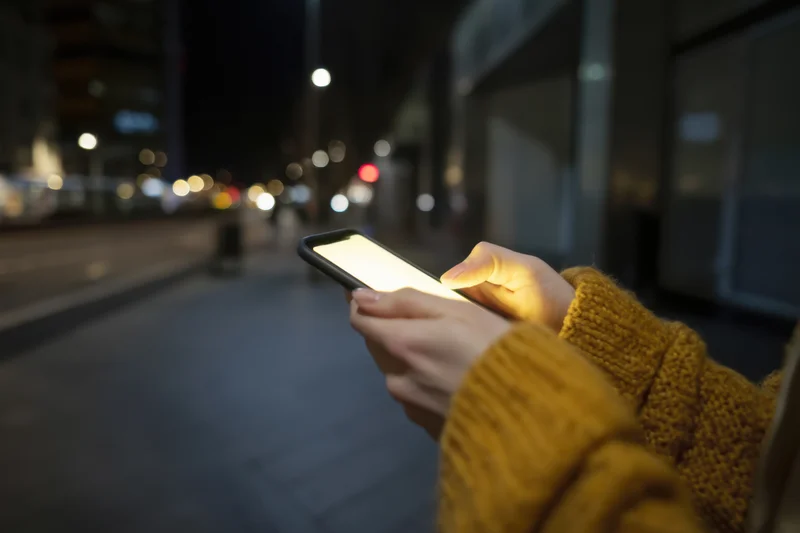 Hands holding smartphone with glowing screen on city sidewalk at night