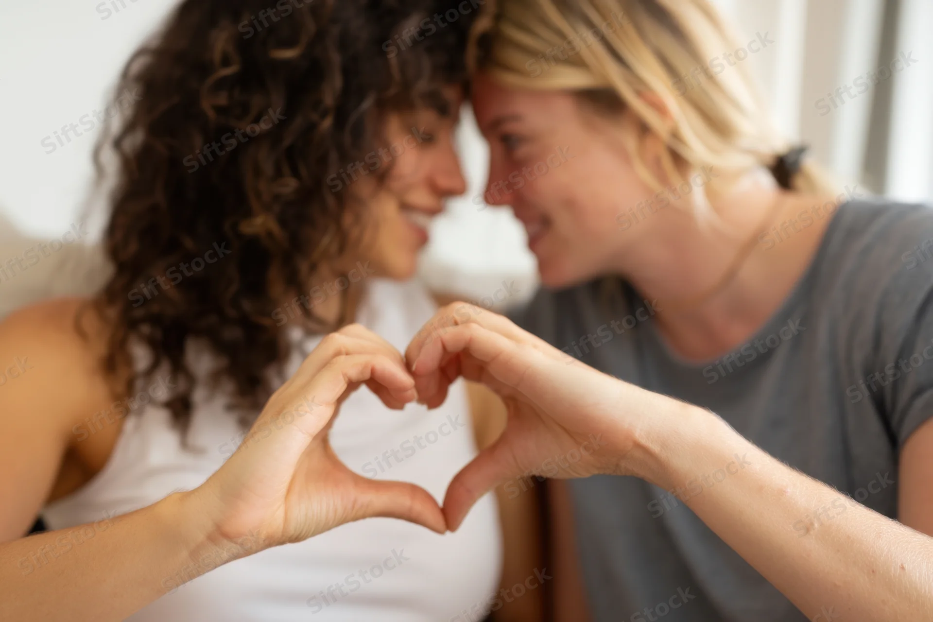 Two women smiling and forming a heart shape with their hands while facing each other indoors