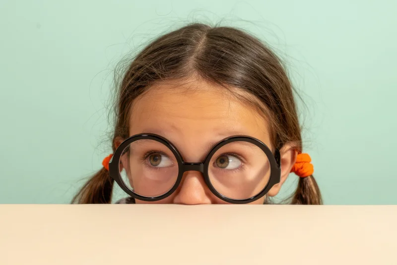 Young girl with oversized round glasses peeking over a beige surface against a mint background