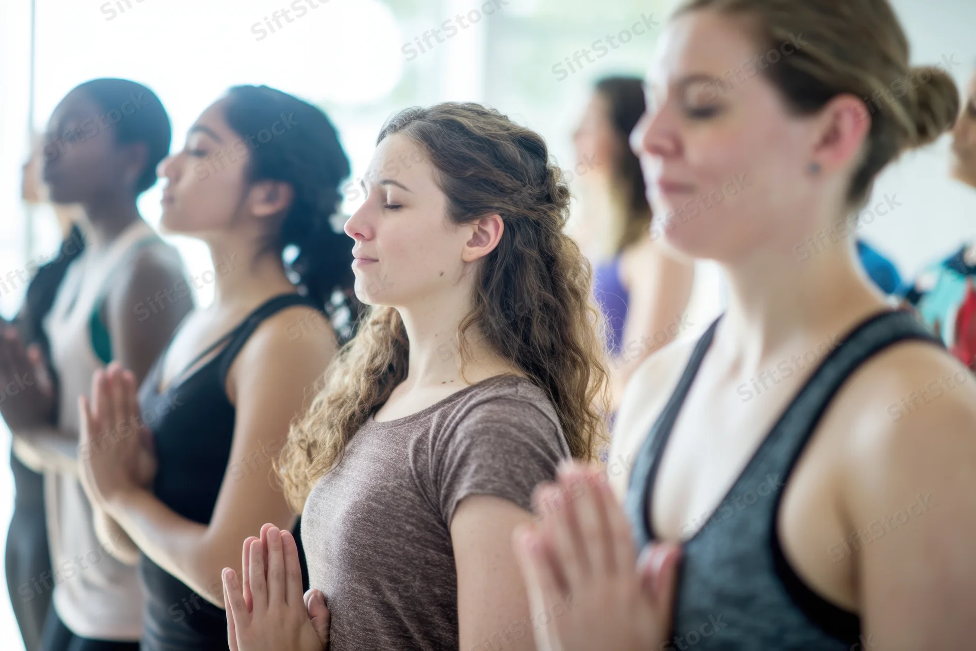 Women practicing yoga in a sunlit studio with hands in prayer position, eyes closed, standing in a row