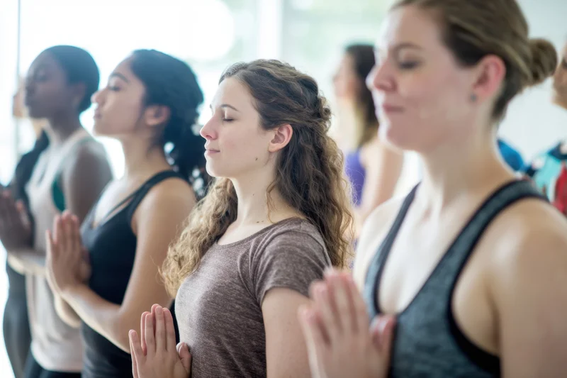 Women practicing yoga in a sunlit studio with hands in prayer position, eyes closed, standing in a row
