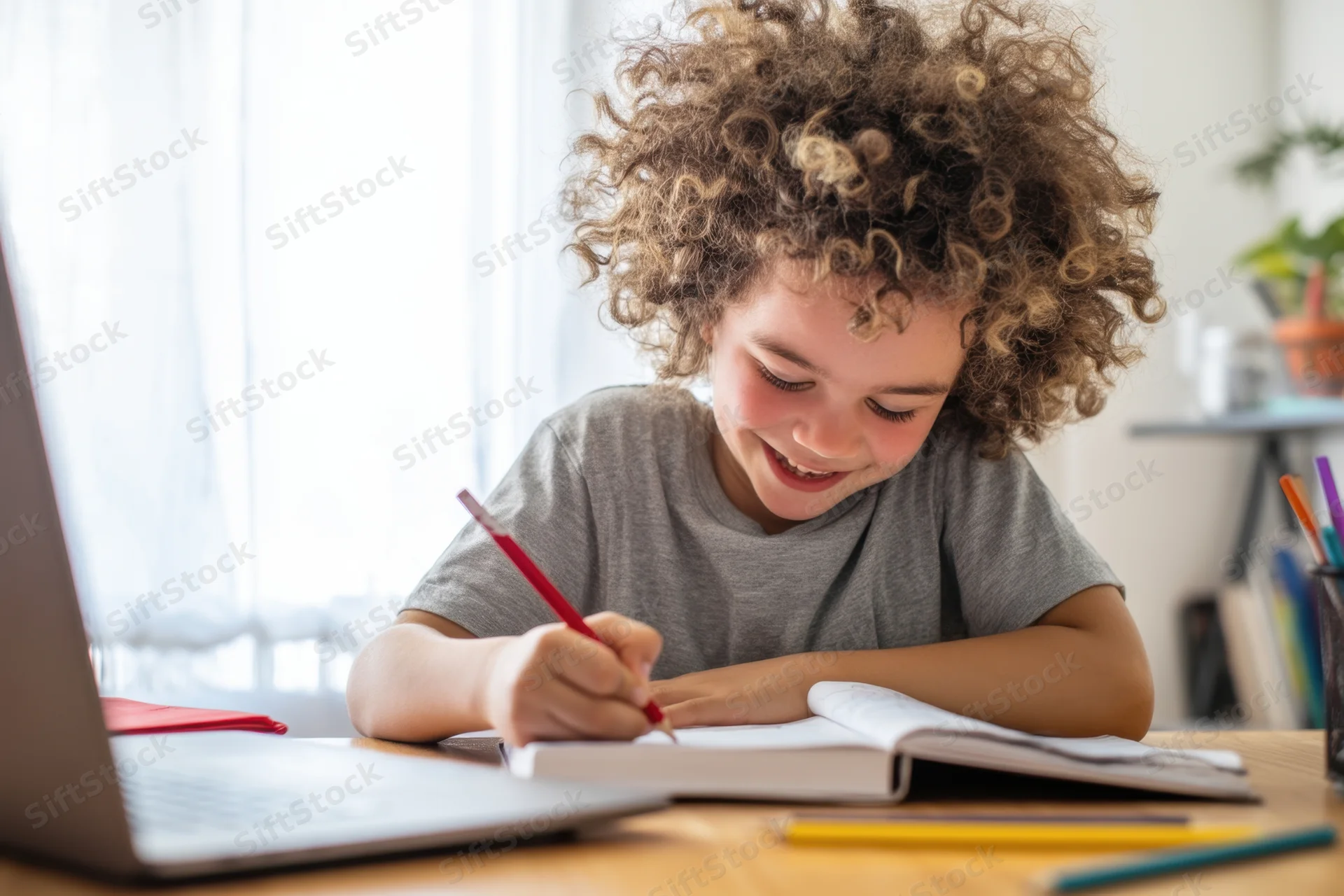 Young girl with curly hair writing in a notebook at a wooden desk beside a laptop
