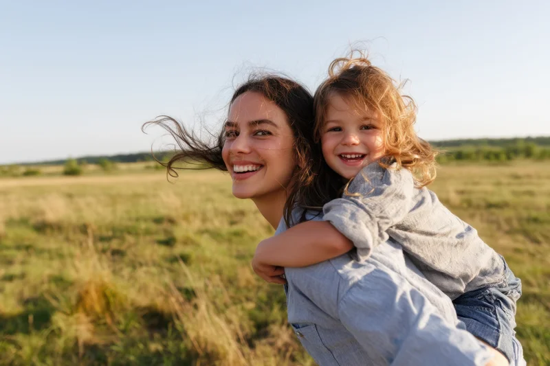 Mother carrying child on her back in a sunlit field, both smiling with wind in their hair