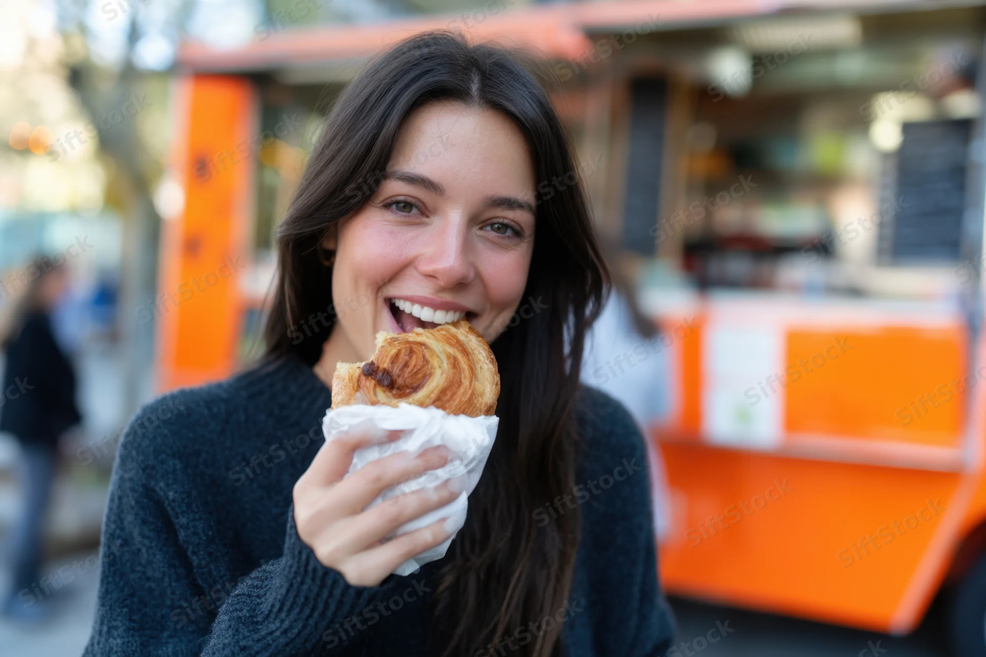 Woman smiling while eating a croissant wrapped in paper in front of a food truck