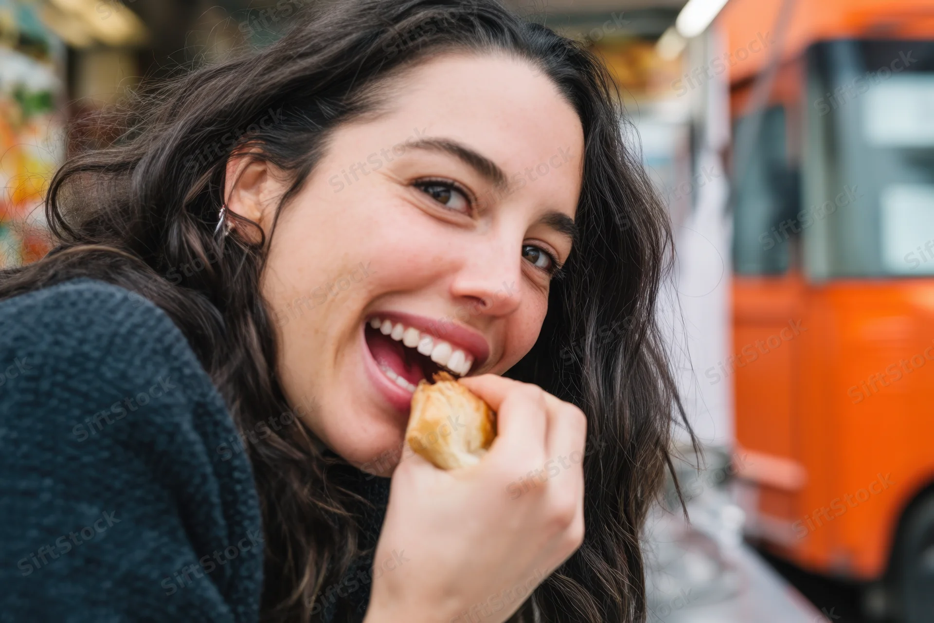 Woman biting into a pastry while smiling in front of an orange food truck