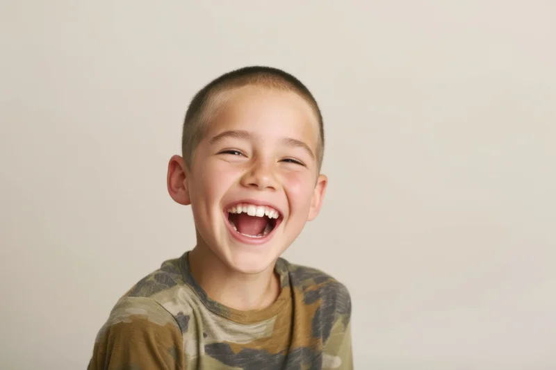 Young boy laughing with wide open mouth and crinkled eyes, wearing camouflage tshirt against light beige background