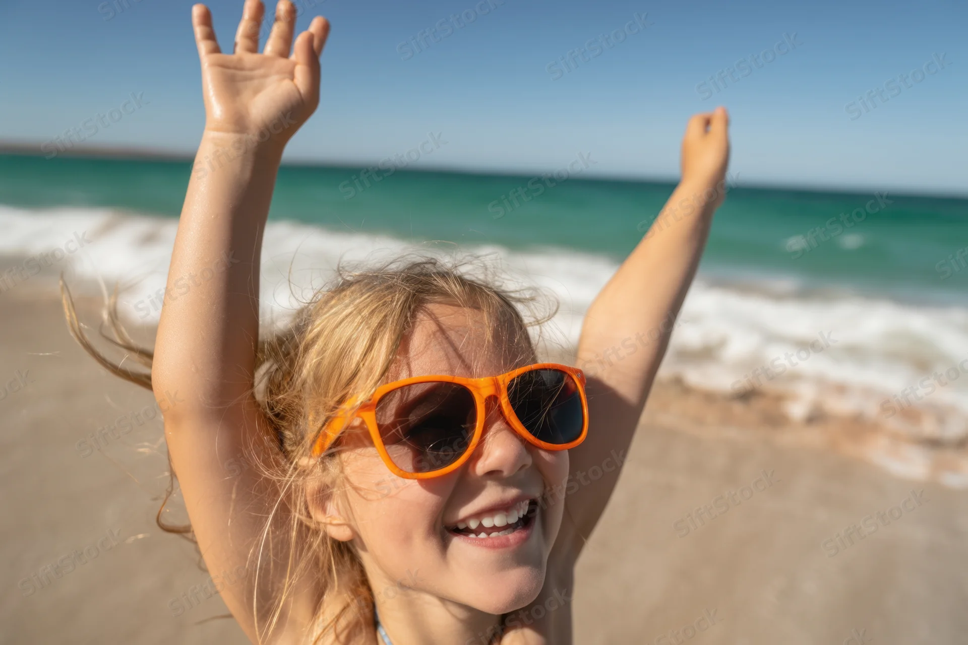 Young girl with orange sunglasses smiling with arms raised on sandy beach by ocean