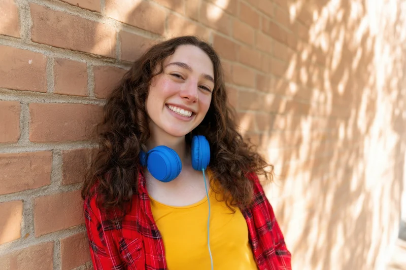 Young woman smiling with blue headphones around neck leaning against sunlit brick wall