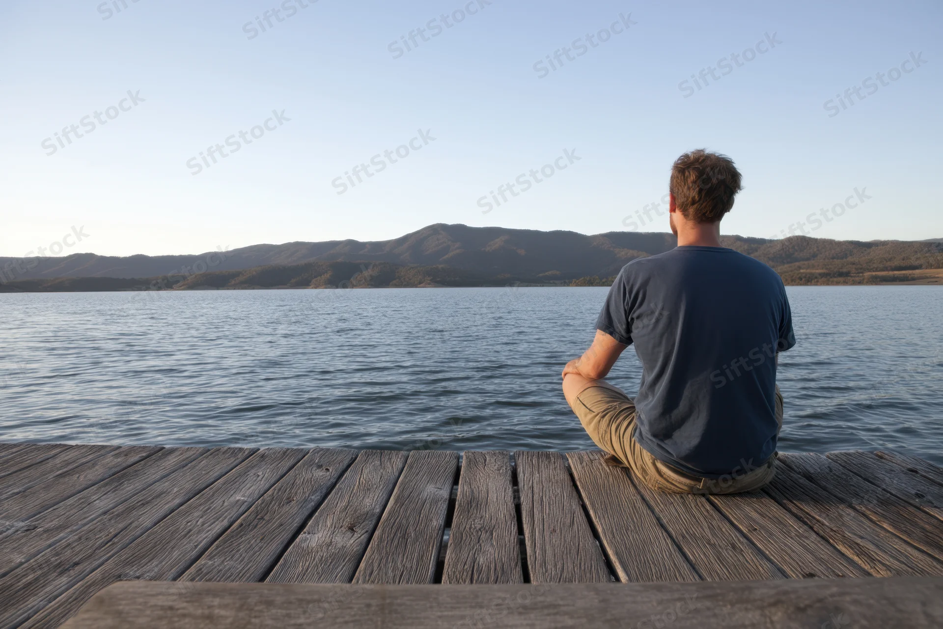 Man sitting cross-legged on wooden dock facing lake and mountains