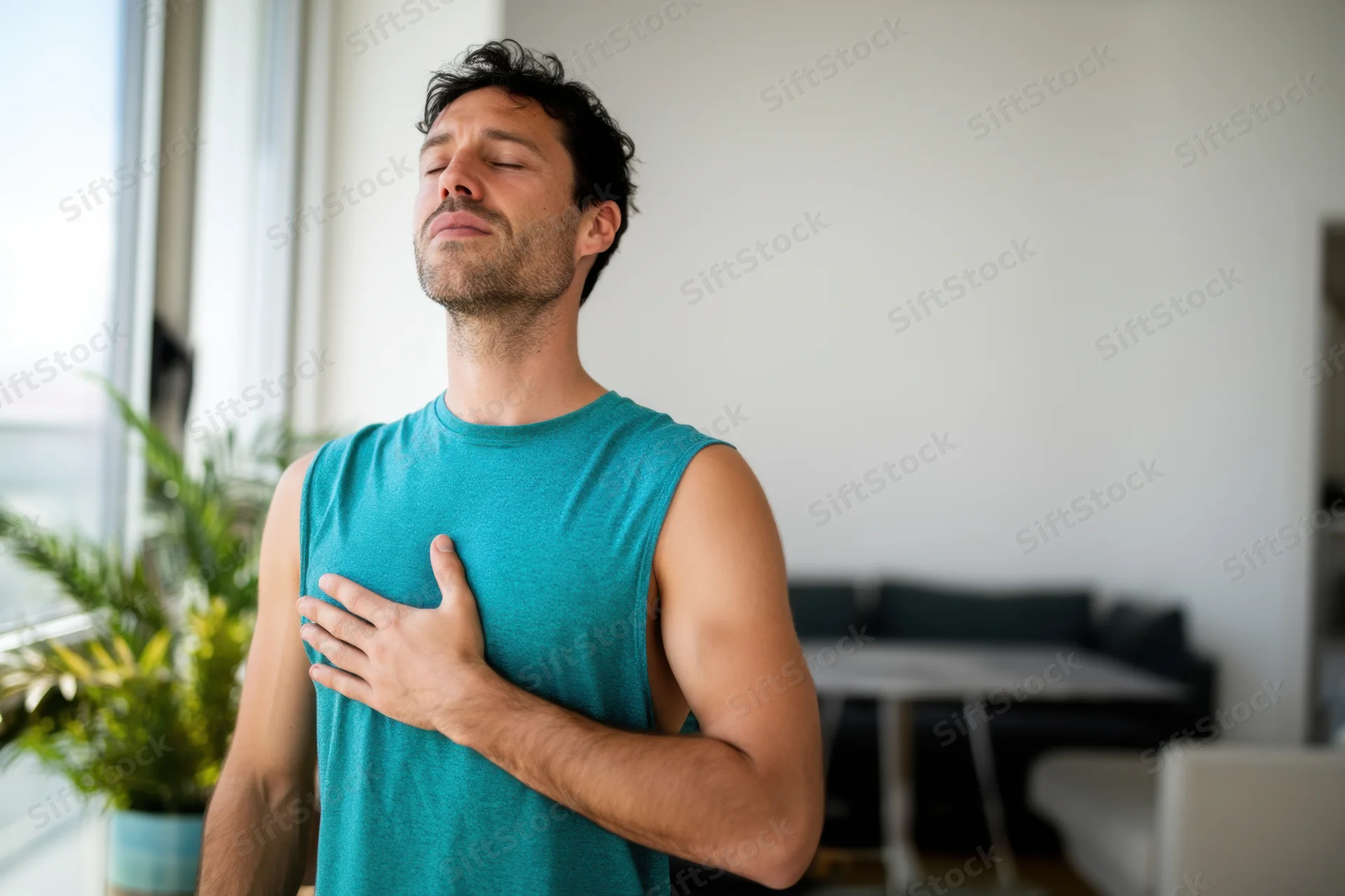 Man meditating with hand on chest in living room near window