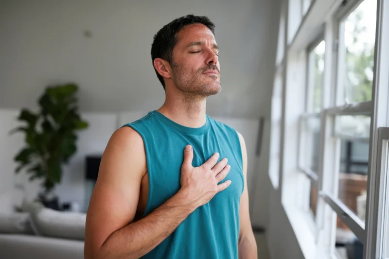 Man meditating with hand on chest near window in living room