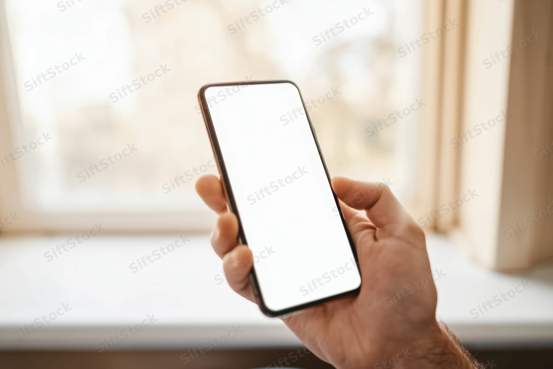 Hand holding smartphone with blank white screen near window indoors