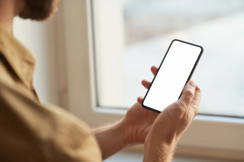 Man holding smartphone with blank white screen near window indoors