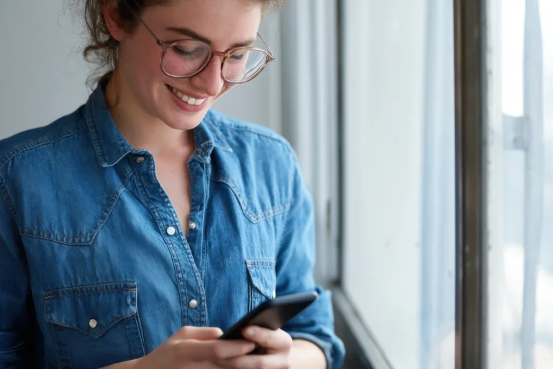 Woman smiling while using smartphone by window in denim shirt