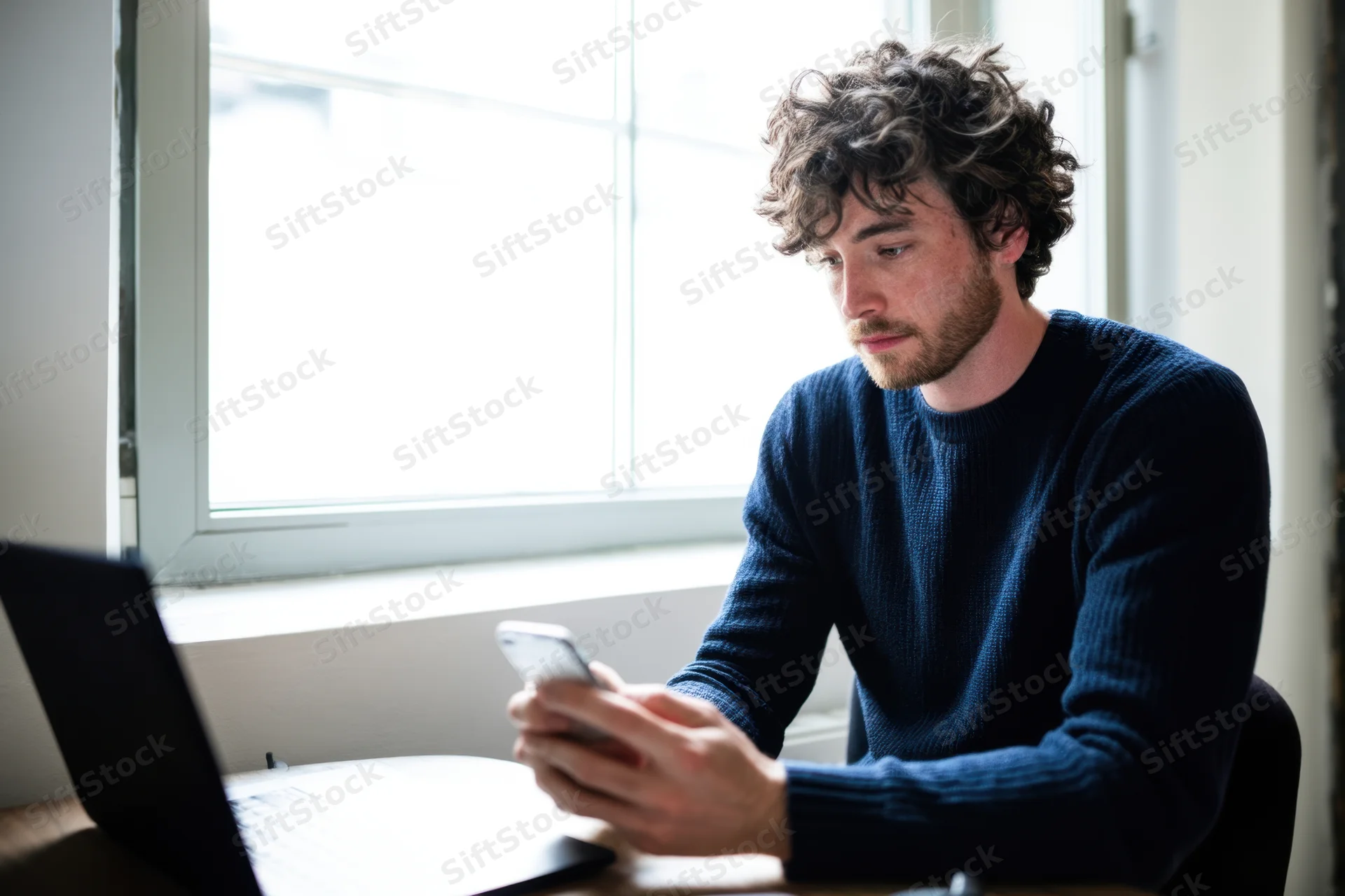 Man in dark blue sweater using smartphone at desk with laptop nearby by window