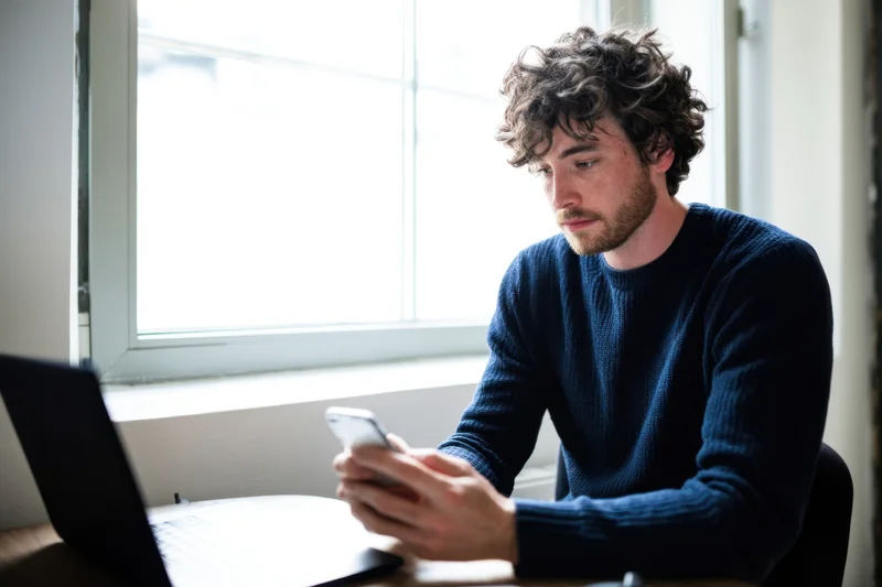Man in dark blue sweater using smartphone at desk with laptop nearby by window