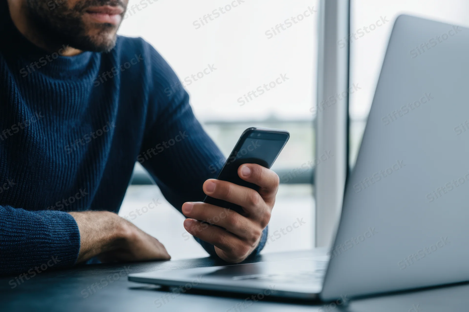 Man holding smartphone while sitting at desk with laptop in office setting