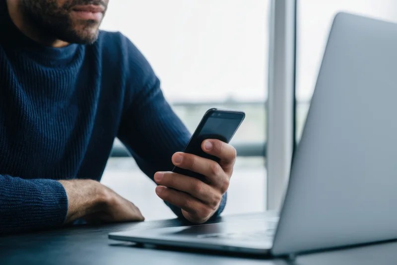 Man holding smartphone while sitting at desk with laptop in office setting