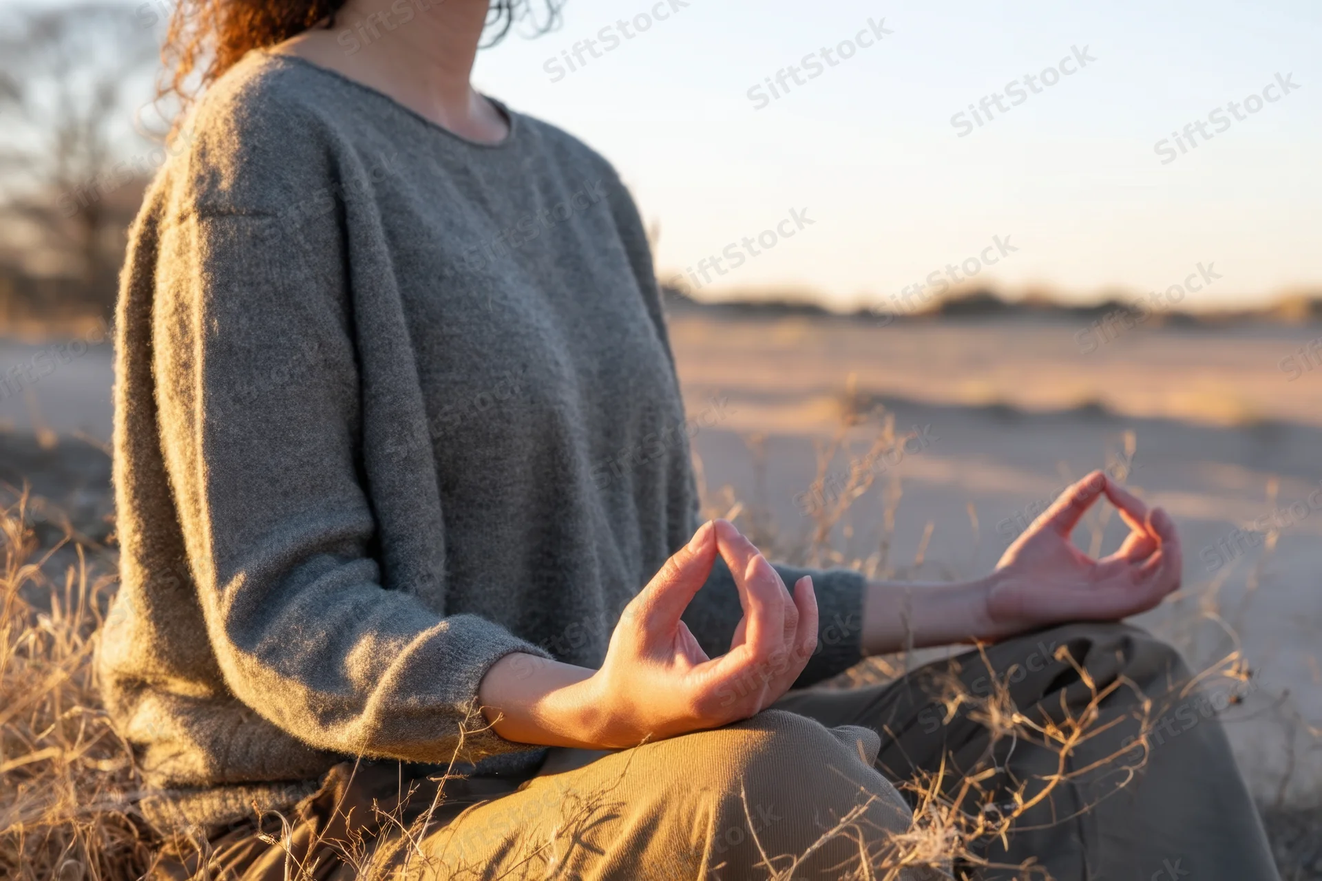 Woman meditating with hands in gyan mudra seated in a dry grass field at sunset