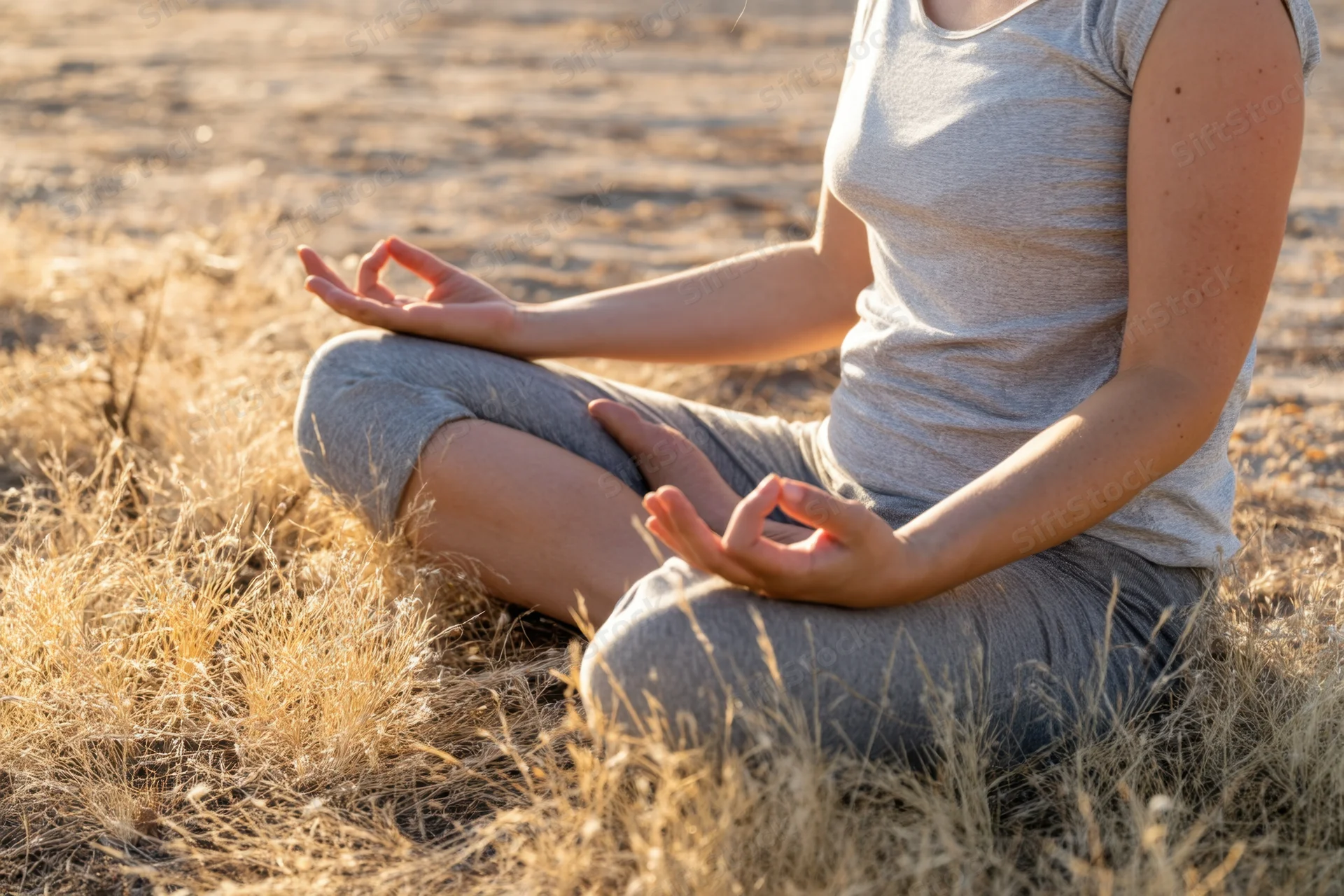 Woman meditating cross-legged in dry grass field with hands in gyan mudra during golden hour