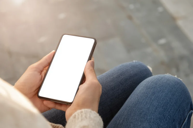 Woman holding smartphone with blank white screen outdoors while sitting on pavement