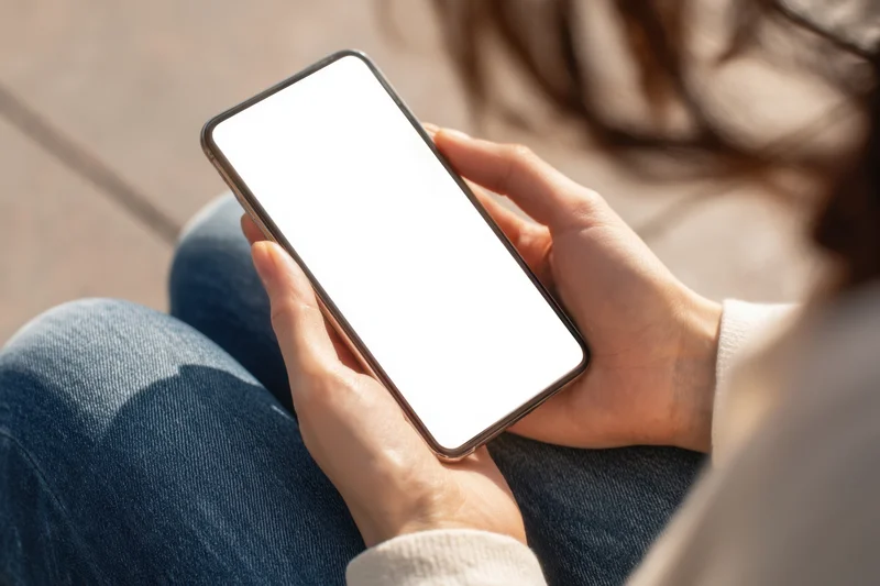 Woman holding smartphone with blank white screen while sitting outdoors in casual clothing