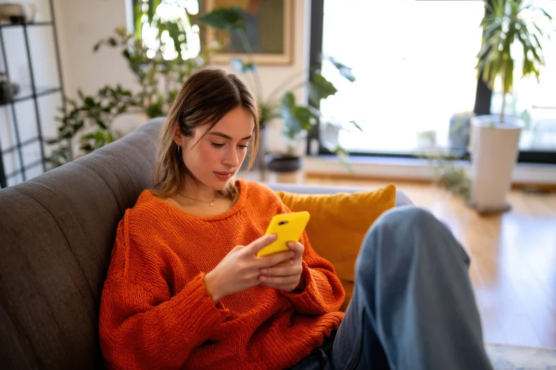 Woman in orange sweater scrolling on smartphone while sitting on sofa in living room