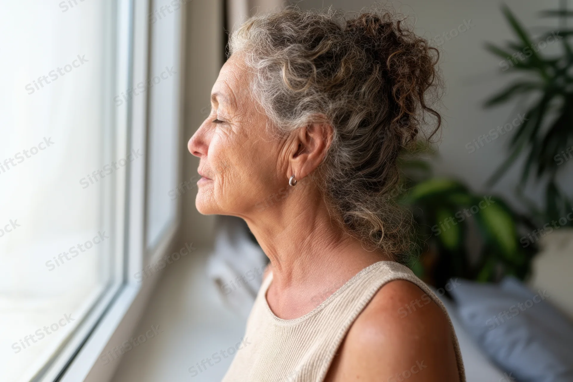 Senior woman in profile with eyes closed looking out window in bedroom