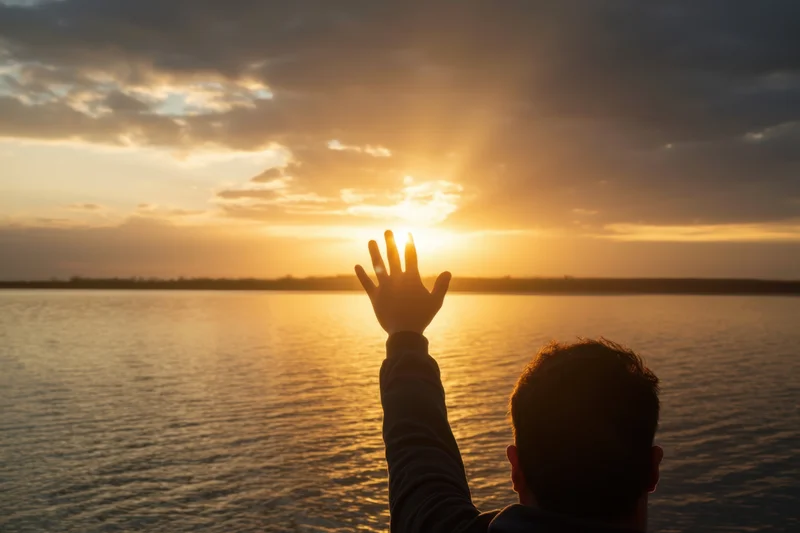 Man reaching hand toward sunset over lake