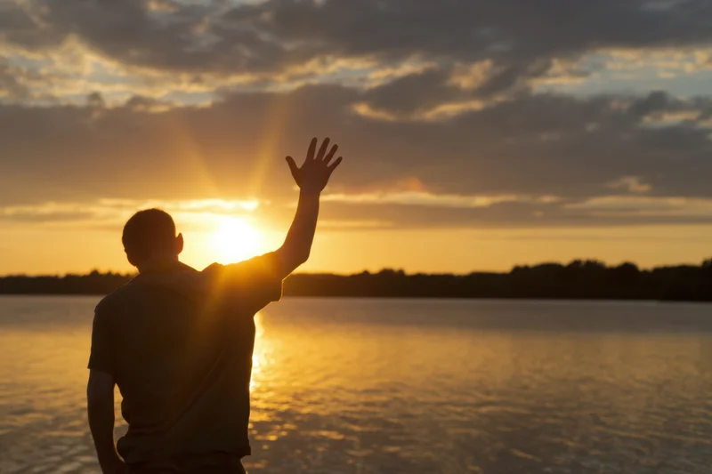 Man in silhouette reaching toward sunset over a lake