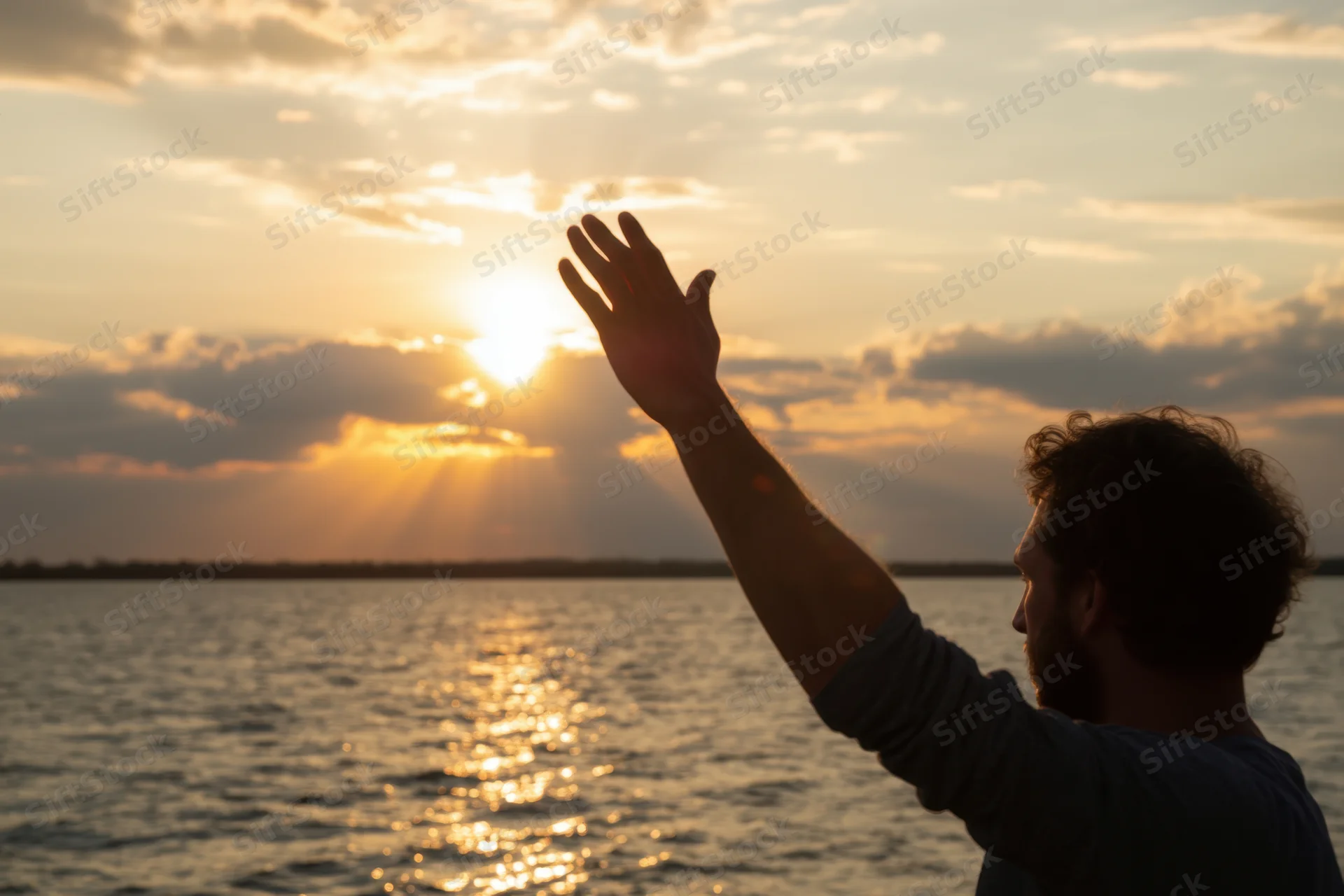 Man in silhouette reaching toward the sun over the ocean at sunset