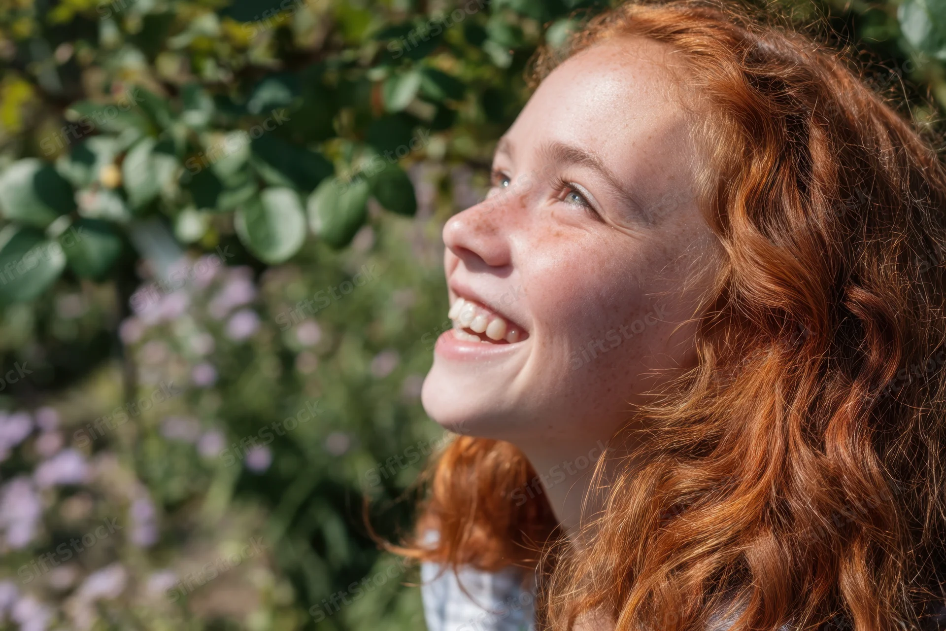Smiling red-haired girl with freckles in profile outdoors in a garden with green foliage and purple flowers
