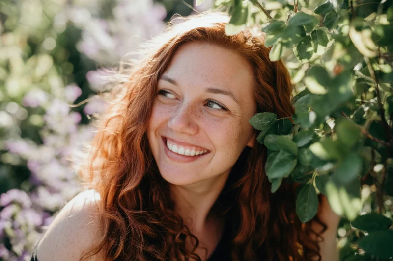 Young woman with red hair and freckles smiling amidst green foliage in a garden