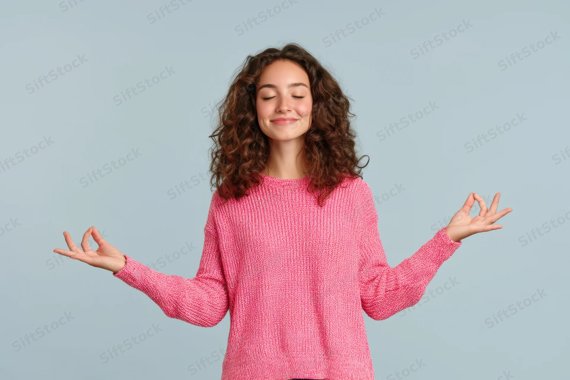 Woman meditating with eyes closed and hands in yoga gesture wearing pink knit sweater against light blue background