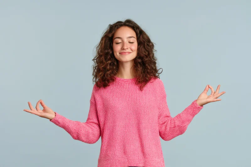 Woman meditating with eyes closed and hands in yoga gesture wearing pink knit sweater against light blue background