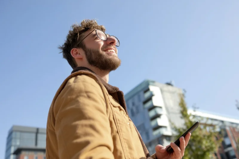 Smiling young man in tan jacket holding smartphone outdoors in urban setting
