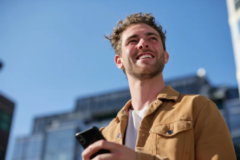 Young man smiling while holding smartphone outdoors in urban setting