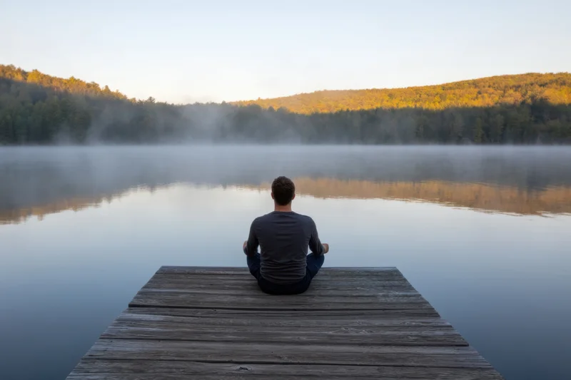 Man meditating cross-legged on wooden dock facing misty lake at sunrise