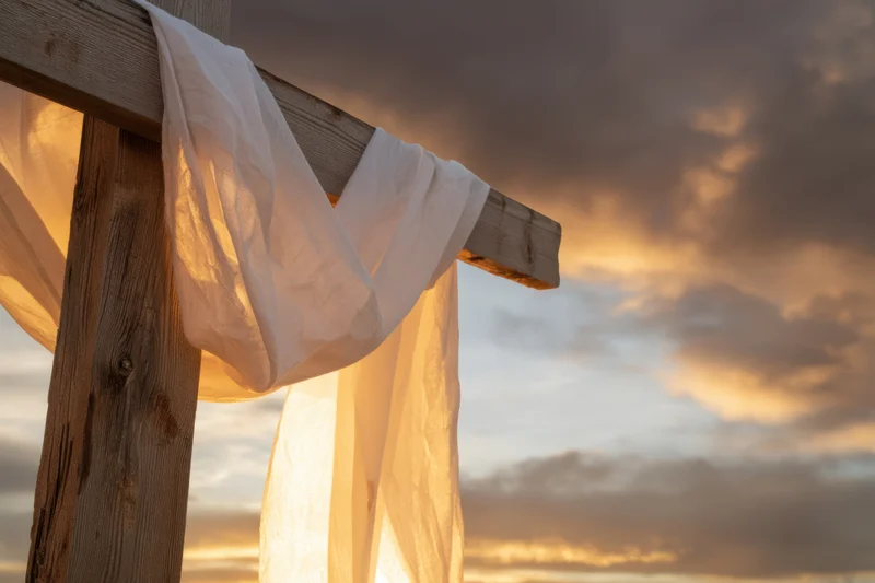 Weathered wooden cross with white fabric draped over it against a dramatic sunset sky