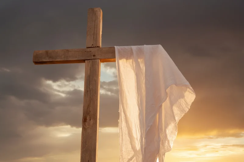 Weathered wooden cross with white cloth draped over it against dramatic sunset sky
