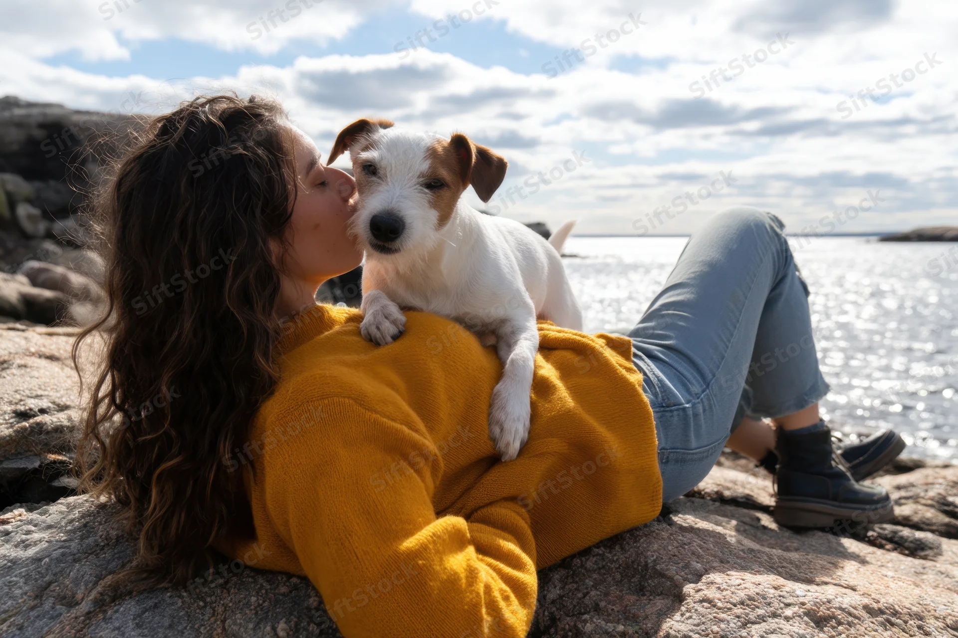 Woman lying on rocky waterfront kissing jack russell dog