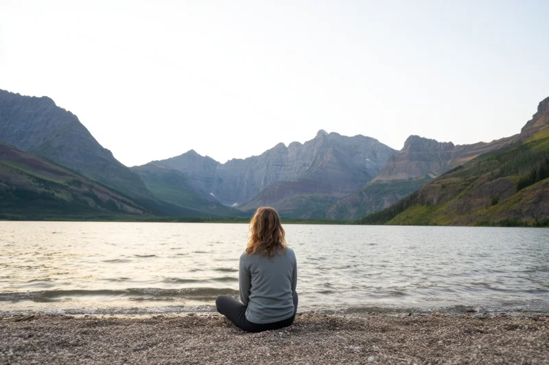 Woman sitting cross-legged on pebbled shore facing lake and mountains